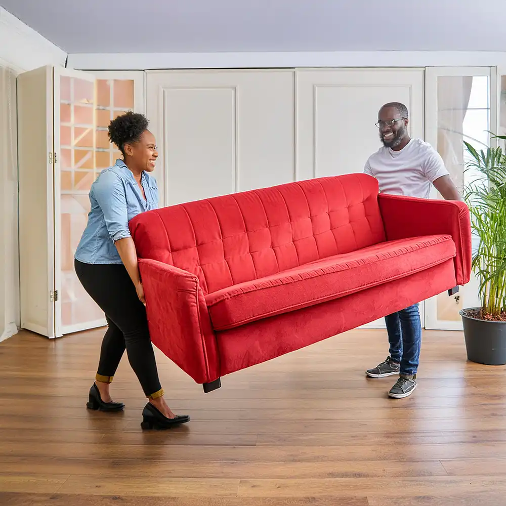 A smiling middle-aged couple moving a large red couch into an empty, large open space room with hardwood floors and white paneled walls.)