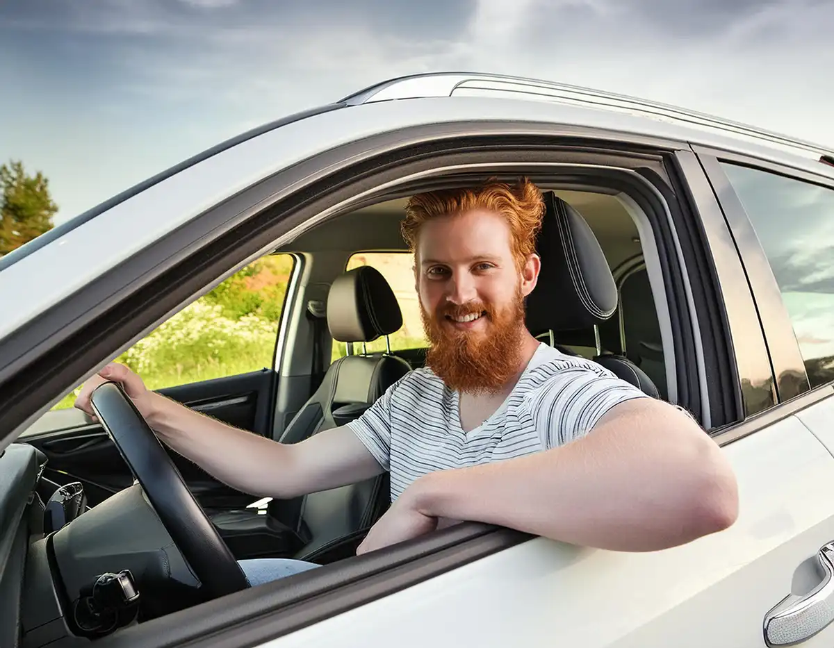 A smiling young, red-headed man with a beard driving a white SUV. He has one hand on the steering wheel and his other arm resting in the open window.